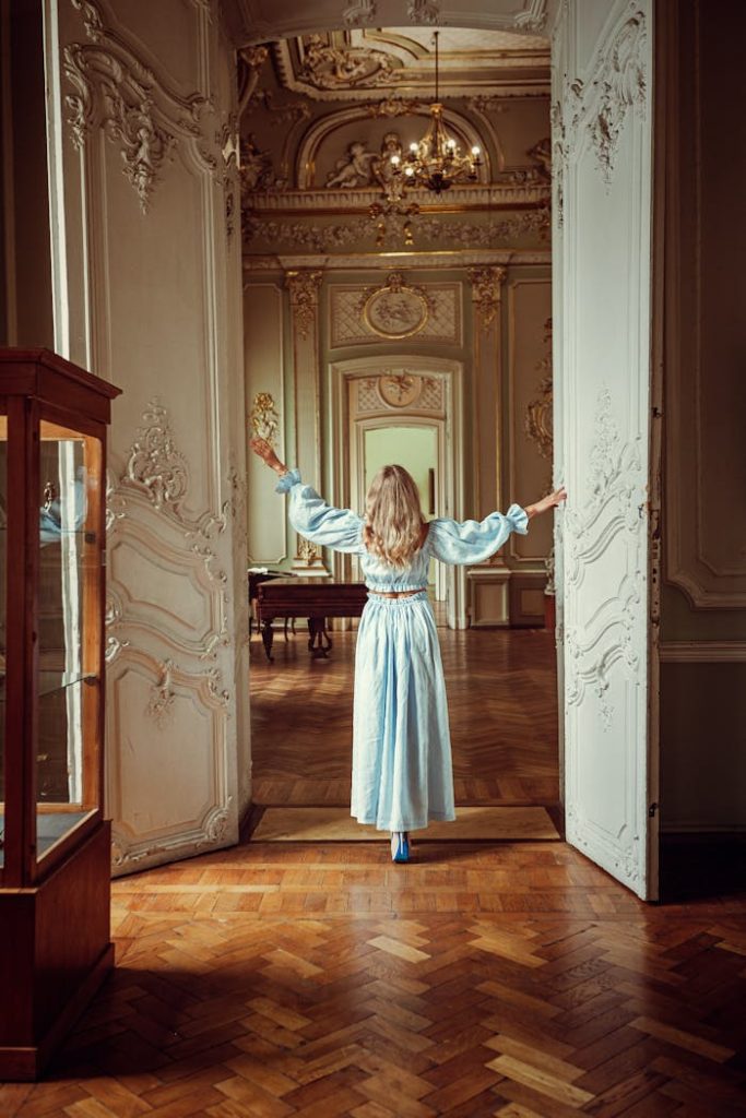 a-back-view-of-a-woman-in-light-blue-terno-clothes-9620214 A woman in a light blue dress stands in an ornately decorated room, arms outstretched.